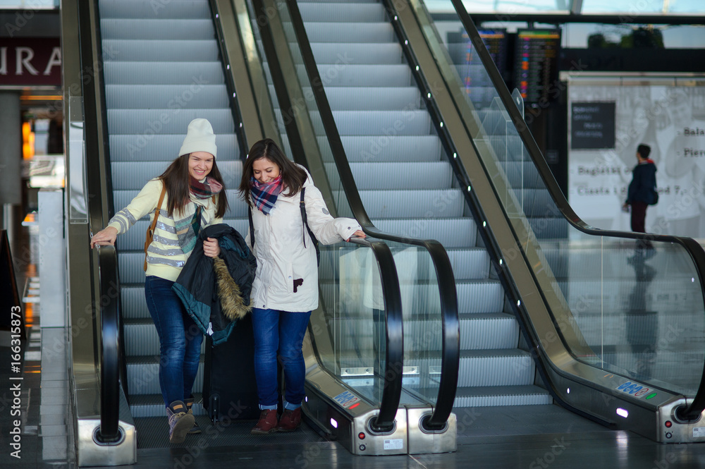 Fototapeta premium Two young women with small luggage on the escalator at the airport.