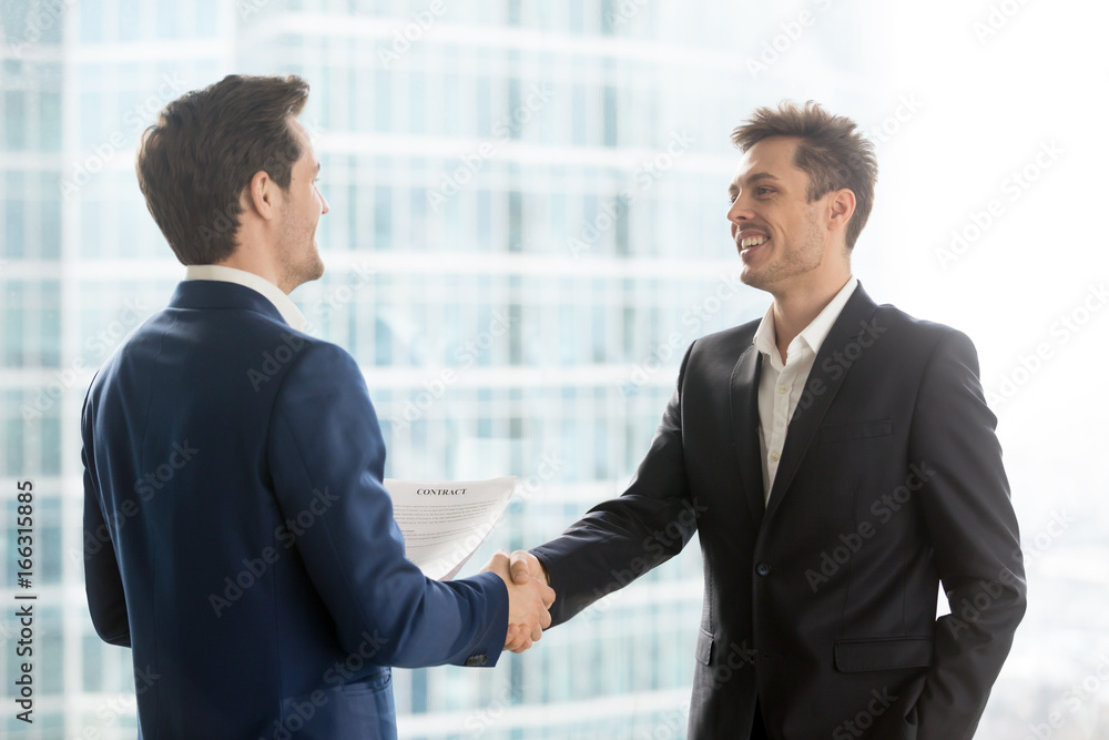 Happy satisfied business partners shaking hands after concluding contract for services on city building background, two smiling businessmen seal good deal, binding bargain with firm strong handshake Happy satisfied business partners shaking hands after concluding contract for services on city building background, two smiling businessmen seal good deal, binding bargain with firm strong handshake