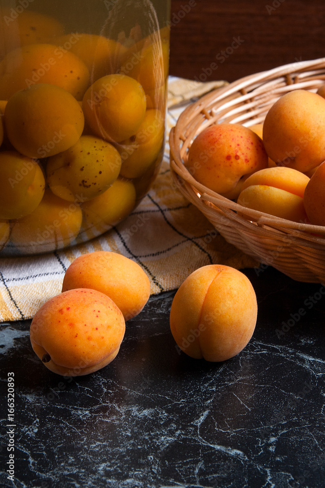 Fresh organic harvested apricots in basket and big glass jar with canned apricot compote on dark marble.. Fresh organic harvested apricots in basket and big glass jar with canned apricot compote on dark marble..