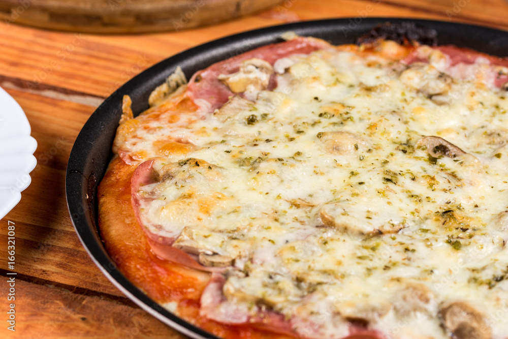 Baked pizza with ham mushrooms grated cheese oregano on the round baking pan on the table Baked pizza with ham mushrooms grated cheese oregano on the round baking pan on the table