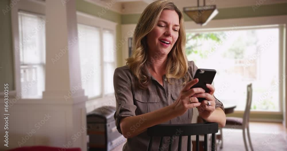 Woman browsing photos on smartphone in dining room, seated in chair Woman browsing photos on smartphone in dining room, seated in chair