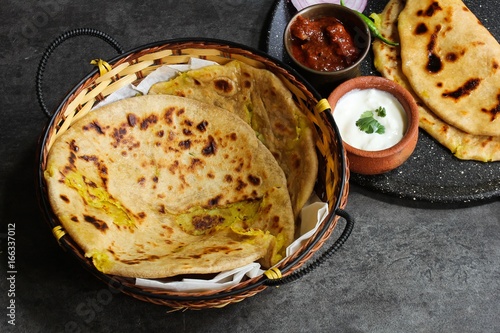 Alu paratha - North Indian Potato stuffed bread served in a basket with raita and mango pickle on dark moody grey background photo