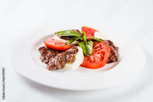 Meat salad. Fried beef with mozzarella, tomato and basil on a white plate on a light background. Close