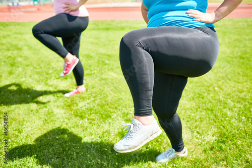Fat woman bending her knee while raising leg during training on green lawn
