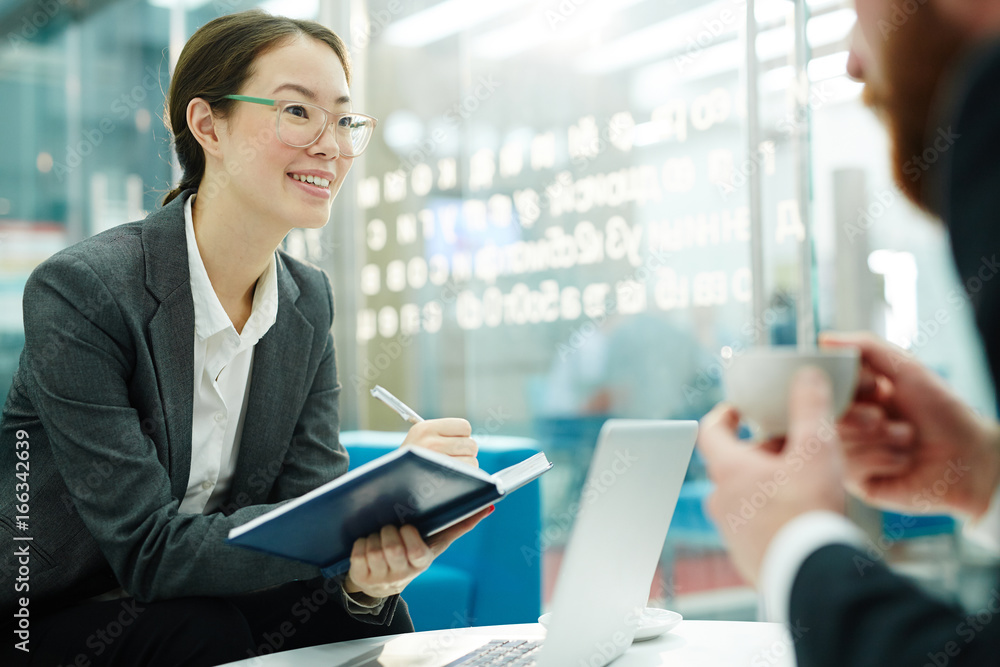 Portrait of smiling Asian business manager taking notes while talking to client during meeting in office