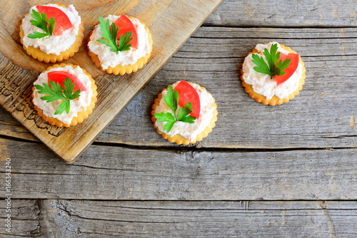 Canape cooked from salty crackers with spicy soft cheese, tomato slice and green parsley leaf. Spicy cheese canape on an old wooden background. Quick aperitif recipe. Top view
