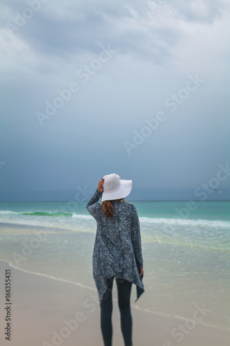 A woman wearing a hat standing on an empty beach with dark clouds. photo