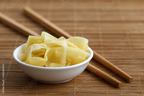 Pickled ginger slices in white ceramic bowl on bamboo mat next to chopsticks.