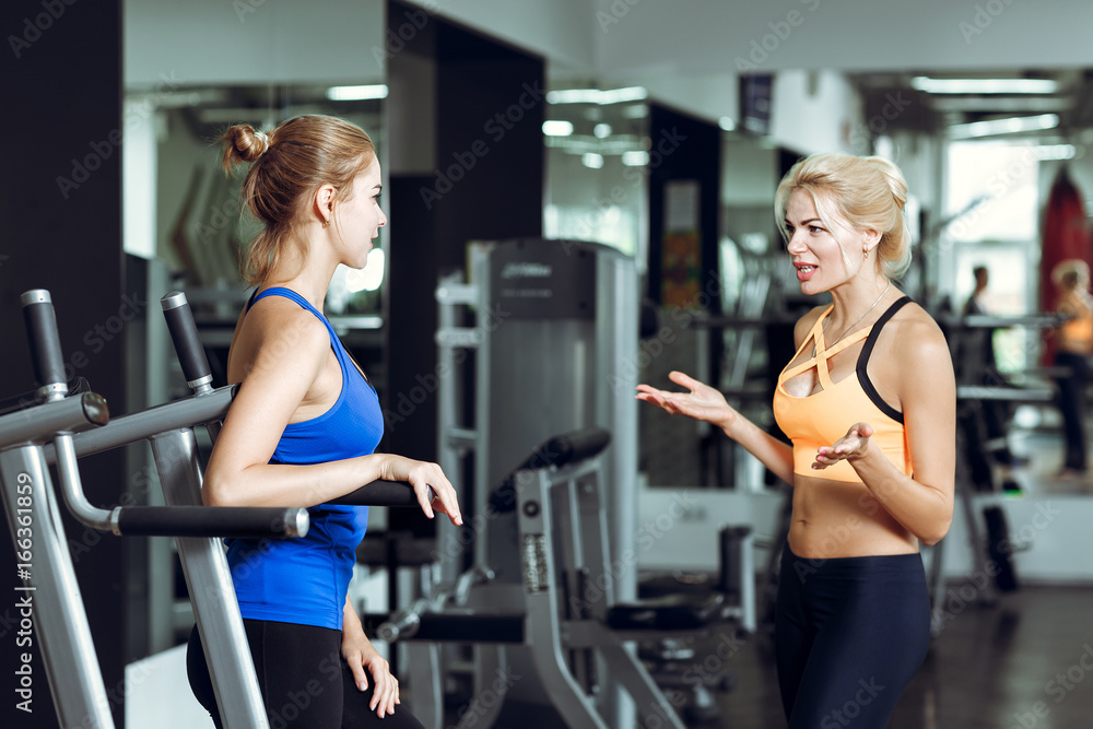 Two athletic blond women talking in gym. Girl communicates with trainer Two athletic blond women talking in gym. Girl communicates with trainer