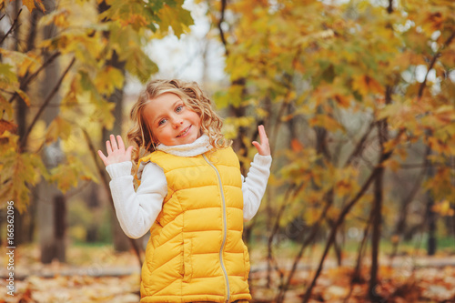 cute toddler girl playing with leaves in autumn park on the walk, wearing fashion yellow outfit