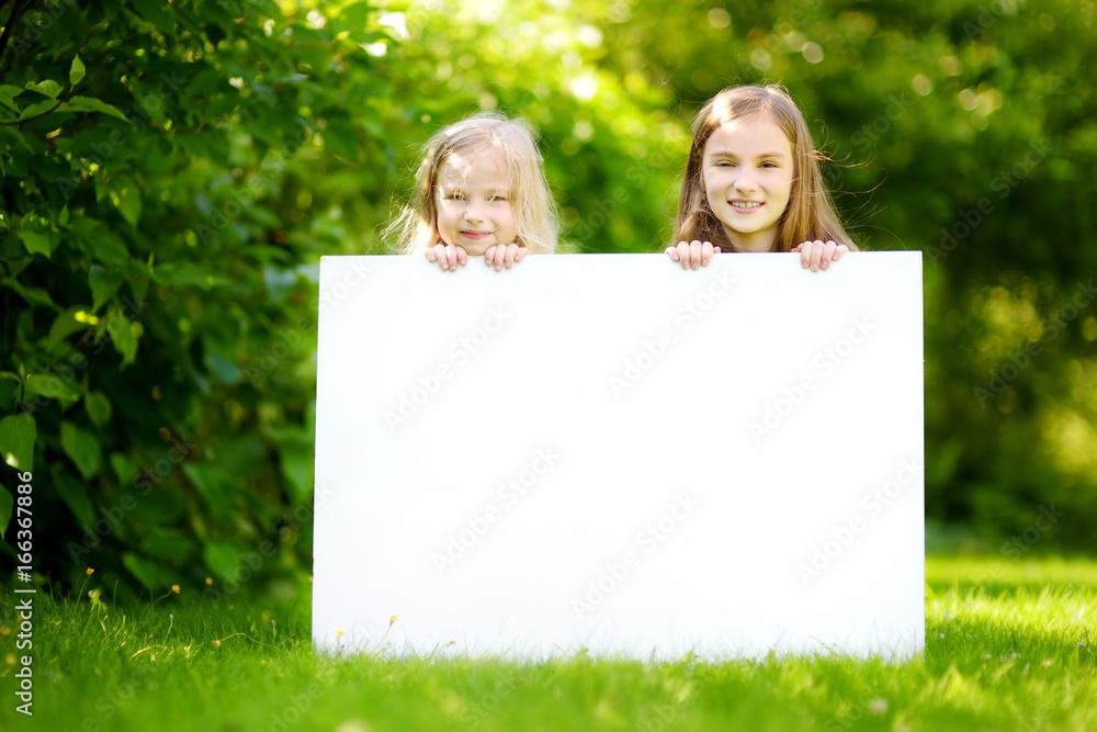 Two cute little sisters holding big blank whiteboard on sunny summer day outdoors Two cute little sisters holding big blank whiteboard on sunny summer day outdoors