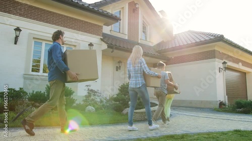 Young exaciting family moving to the new modern light home with big cardboard box, walking throught the garden during the sunset. photo