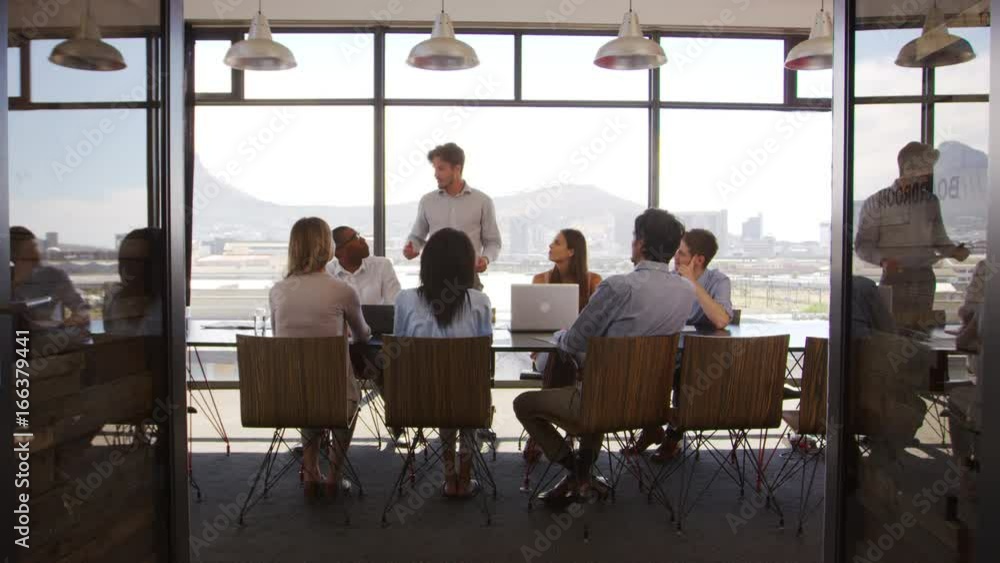 Young man stands addressing colleagues at meeting in a boardroom Young man stands addressing colleagues at meeting in a boardroom