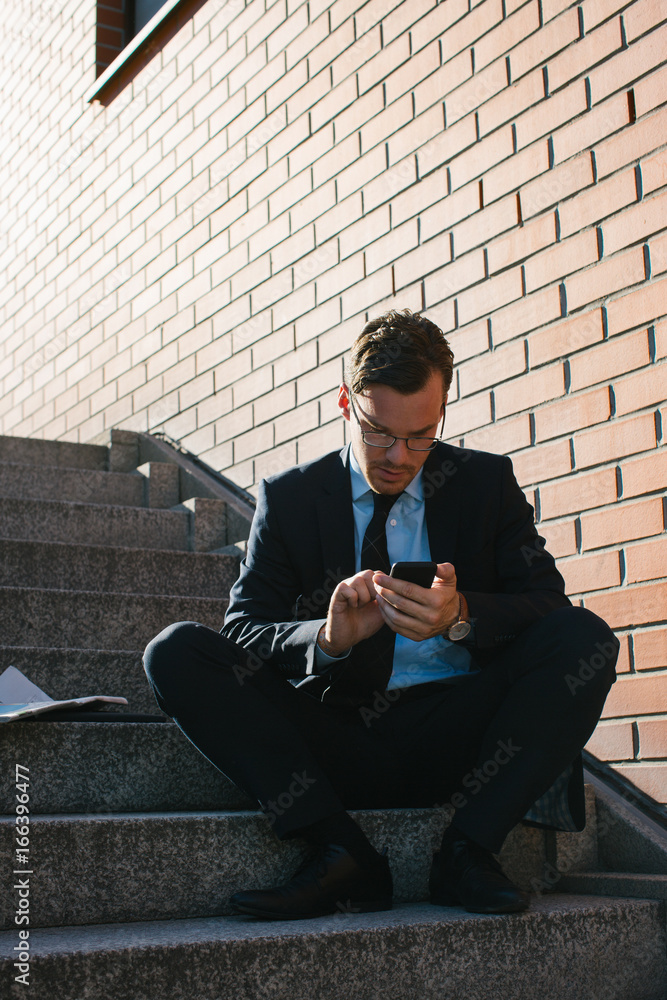 Young Businessman in Blue Suit Sitting on Stairs and Typing on Cellphone Young Businessman in Blue Suit Sitting on Stairs and Typing on Cellphone