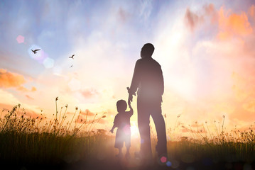 Silhouette of a father holding hands with his young son walking on a grass field at sunrise with birds flying and colorful sky, International Father's Day and family love concept