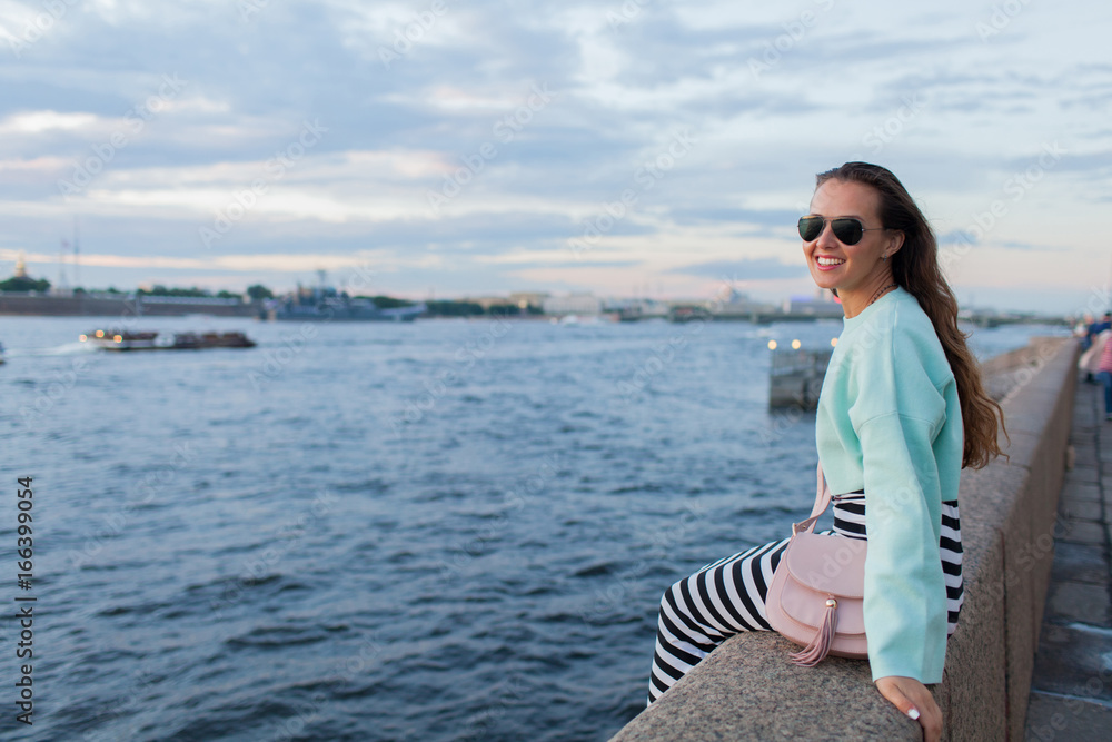 Young and beautiful girl sitting on the embankment of the river. she looks at the sunset and ships passing by. Saint Petersburg, Russia