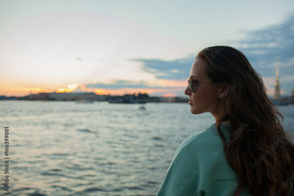 Young and beautiful girl sitting on the embankment of the river. she looks at the sunset and ships passing by. Saint Petersburg, Russia Young and beautiful girl sitting on the embankment of the river. she looks at the sunset and ships passing by. Saint Petersburg, Russia