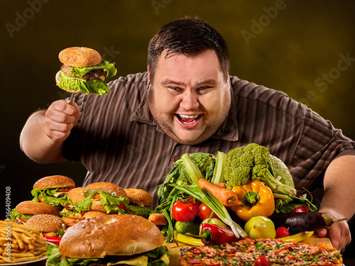 Diet fat man who makes choice between healthy and unhealthy food. Overweight male with hamburgers. Table with food in the foreground.