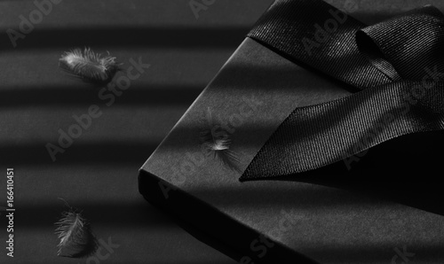 Black gift box on a dark contrasted background, decorated with a textured bow and feathers, creating a romantic atmosphere.
