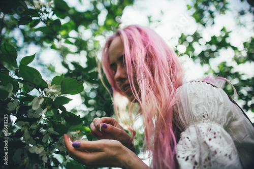 The beautiful girl with pink hair walks among the blossoming apple-tree in the summer