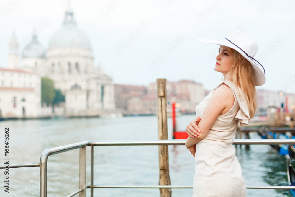 Young female traveler with hat standing on San Marco square with tower and basilica on the background in Venice.