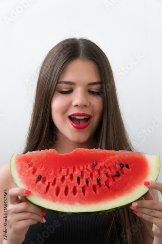 The girl is holding a big watermelon in her hands. The brunette is dressed in a black jersey. The girl stands on a light background