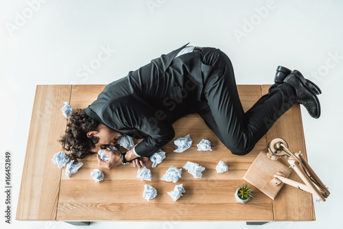 overhead view of depressed businessman lying on table with crumpled documents in office