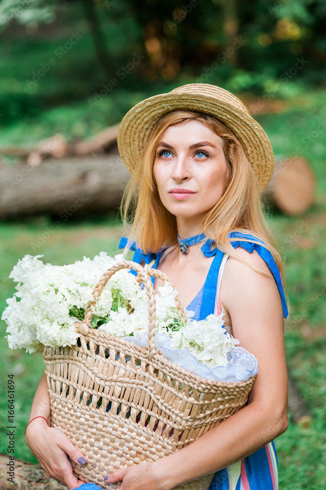 Fototapeta premium Beautiful girl wearing blue dress and hat collect flowers in basket in the wood.