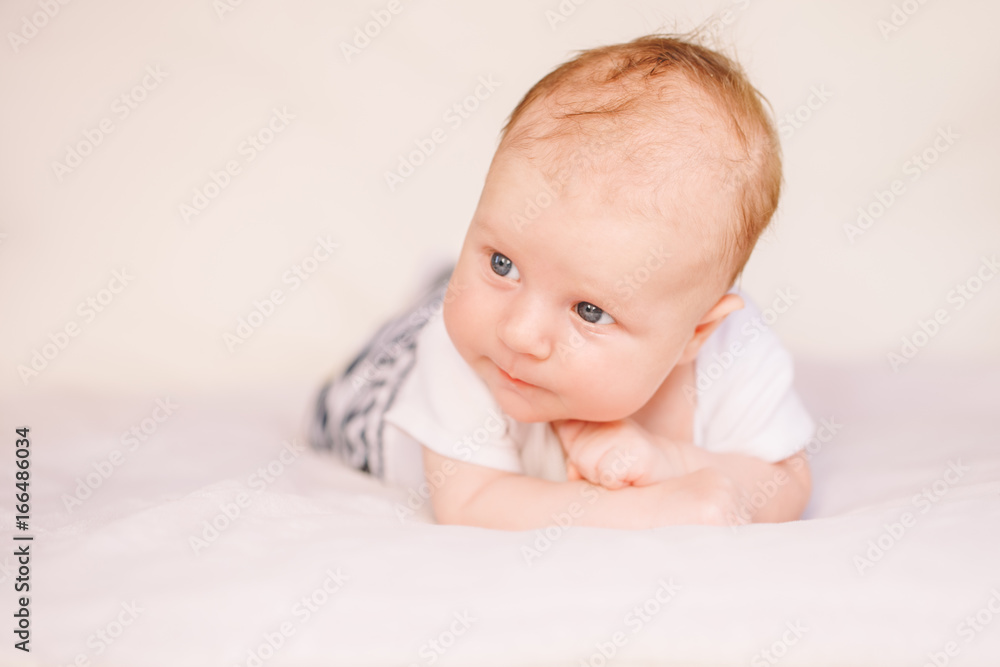Closeup portrait of adorable funny white Caucasian baby with blue grey eyes lying on tummy belly on bed. Aware cute newborn on white light background in studio.