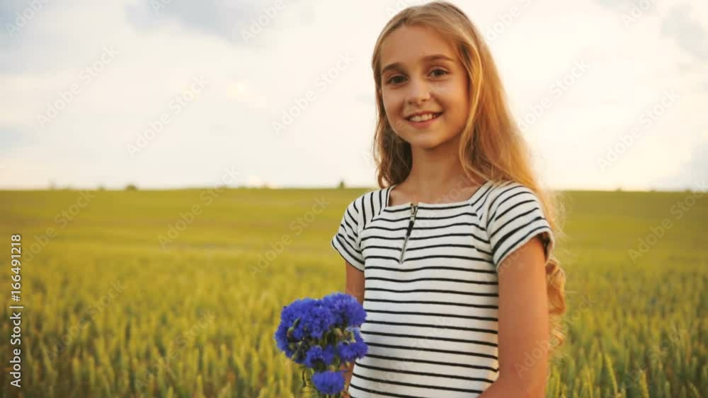 Side portrait of attractive little girl stending in the golden field with beatiful bouquet of cornflowers, turning to the camera and smiling on the camera. Side portrait of attractive little girl stending in the golden field with beatiful bouquet of cornflowers, turning to the camera and smiling on the camera.