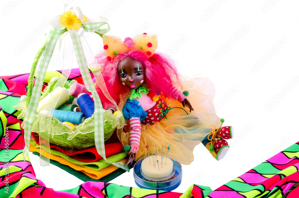 Cute doll sits from above to pieces of colorful cloth, next to it pins, multicoloredcloth  and thread in the basket. Isolated on white background.