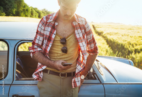 Young Man in 1970s Clothes Leaning Against Blue Vintage Car and Typing on Cellphone photo