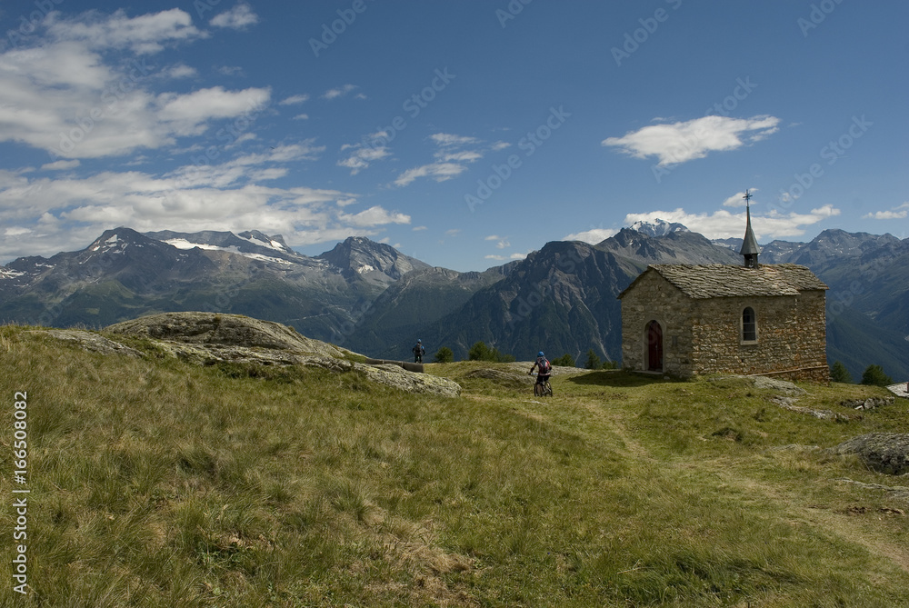 Mountain summer landscape: trail with two electric bicycles E-bike on a sunny day with clouds and a small church in a high mountain village, Belalp, Alps, Switzerland