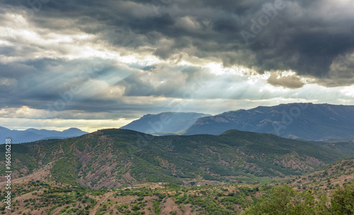 Dramatic stormy sky with sun rays through the clouds over a hilly valley with mountains in the distance