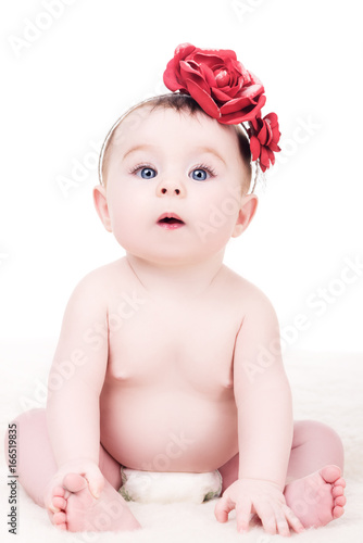 adorable baby girl with headband with a flowers on her head, on white background. surprised child, with big blue eyes, sitting on soft blanket and looking into the camera