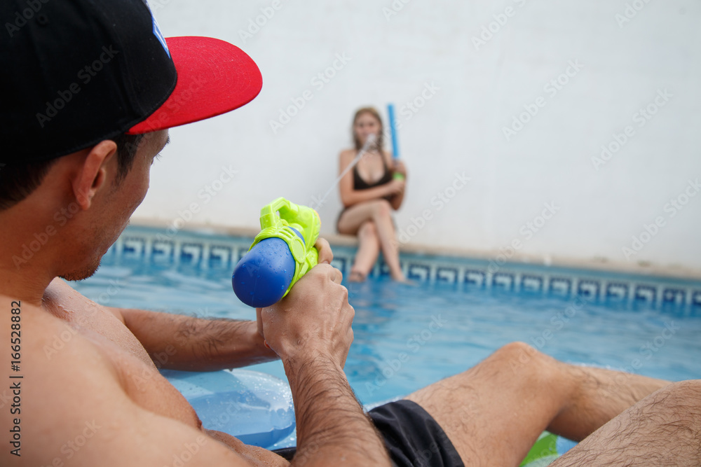 Back view of man on inflatable mat shooting at girl with water gun while relaxing in pool.  