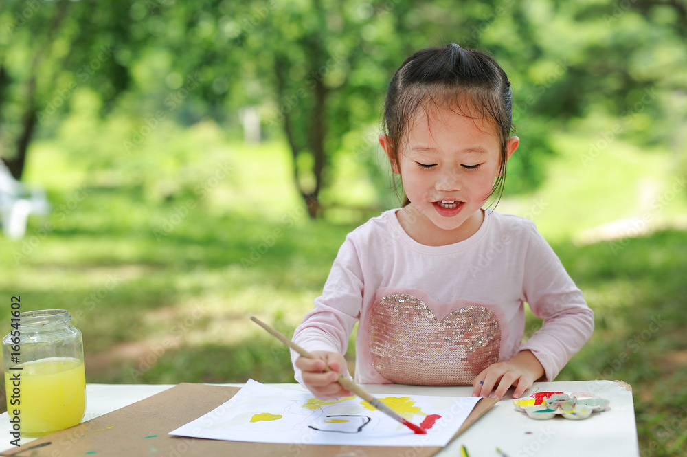 Happy asian child girl sitting at a table in a summer garden painting with paintbrush, Education art concept. Happy asian child girl sitting at a table in a summer garden painting with paintbrush, Education art concept.