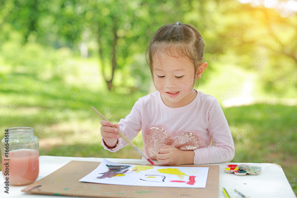Happy asian child girl sitting at a table in a summer garden painting with paintbrush, Education art concept. Happy asian child girl sitting at a table in a summer garden painting with paintbrush, Education art concept.