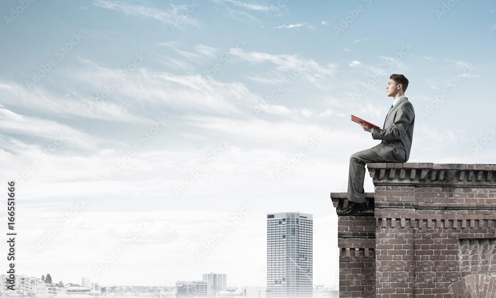 Man on roof edge reading book and cityscape at background Man on roof edge reading book and cityscape at background
