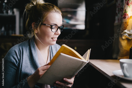beautiful young woman reading interesting book while drinking tea at home, in cafe