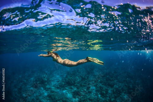 A grirl in yellow bikini and fins snorkeling under water surface in blue sea over coral reef.