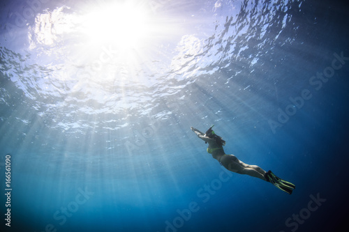 A fridiver girl wearing yellow bikini and fins going up from deep blue to water surface lit with sunlight on background