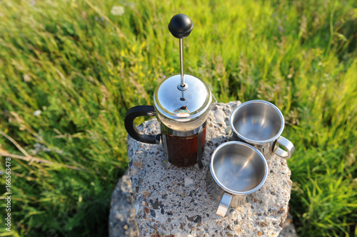 French press with coffee and two metal cups on a stone stand among summer greenery in the open air photo