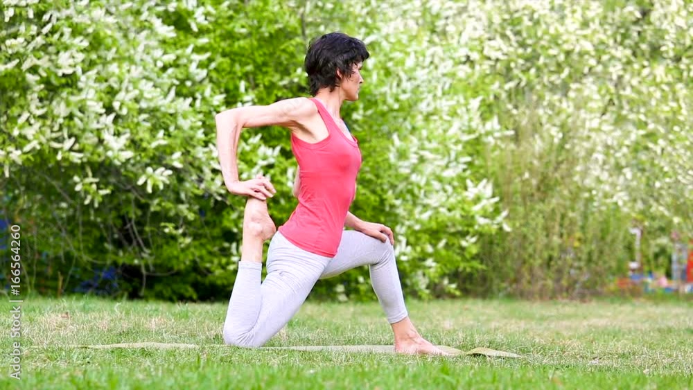 Young brunette woman practices yoga moves on green grass in public place. Slender fit woman doing yoga on exercise mat in city park Young brunette woman practices yoga moves on green grass in public place. Slender fit woman doing yoga on exercise mat in city park