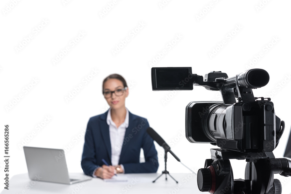 female newscaster sitting at table with laptop and microphone and looking at camera, isolated on white