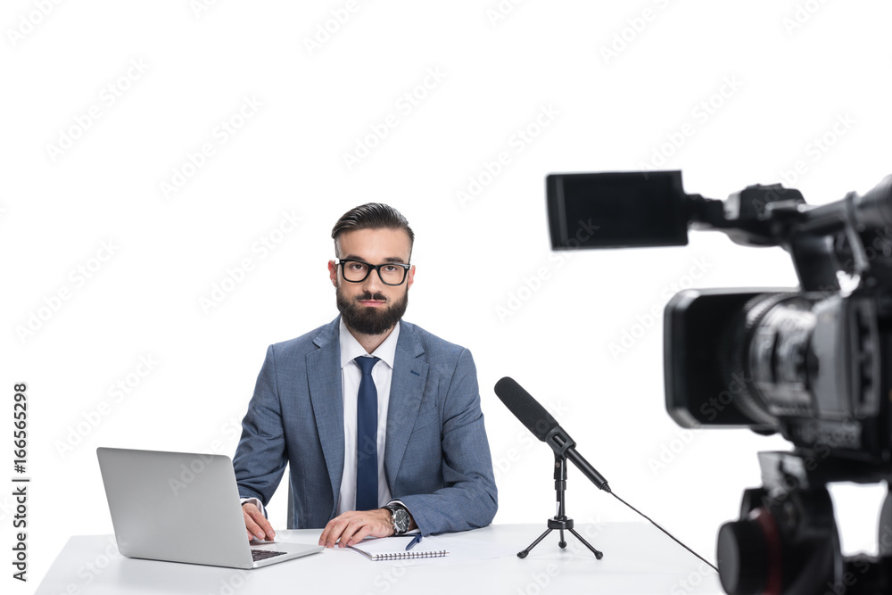 male newscaster sitting at table with laptop, notepad and microphone and looking at camera, isolated on white male newscaster sitting at table with laptop, notepad and microphone and looking at camera, isolated on white