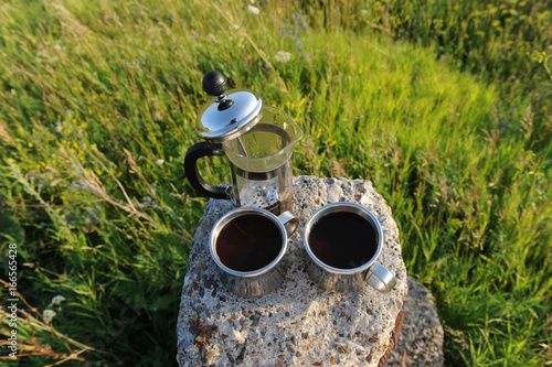 French press with coffee and two metal cups on a stone stand among summer greenery in the open air photo