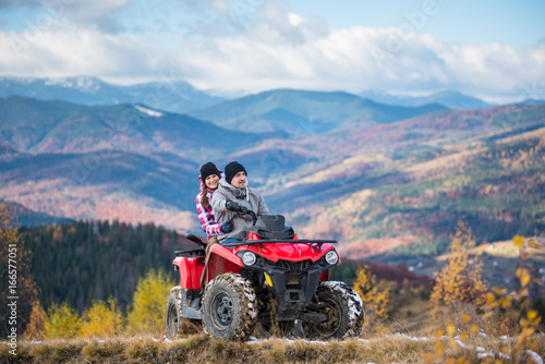 Happy couple in winter sporty clothing on red four-wheeler ATV in mountains, girl sitting behind man and hugging him. Beautiful landscape of mountains and forest at sunny autumn day