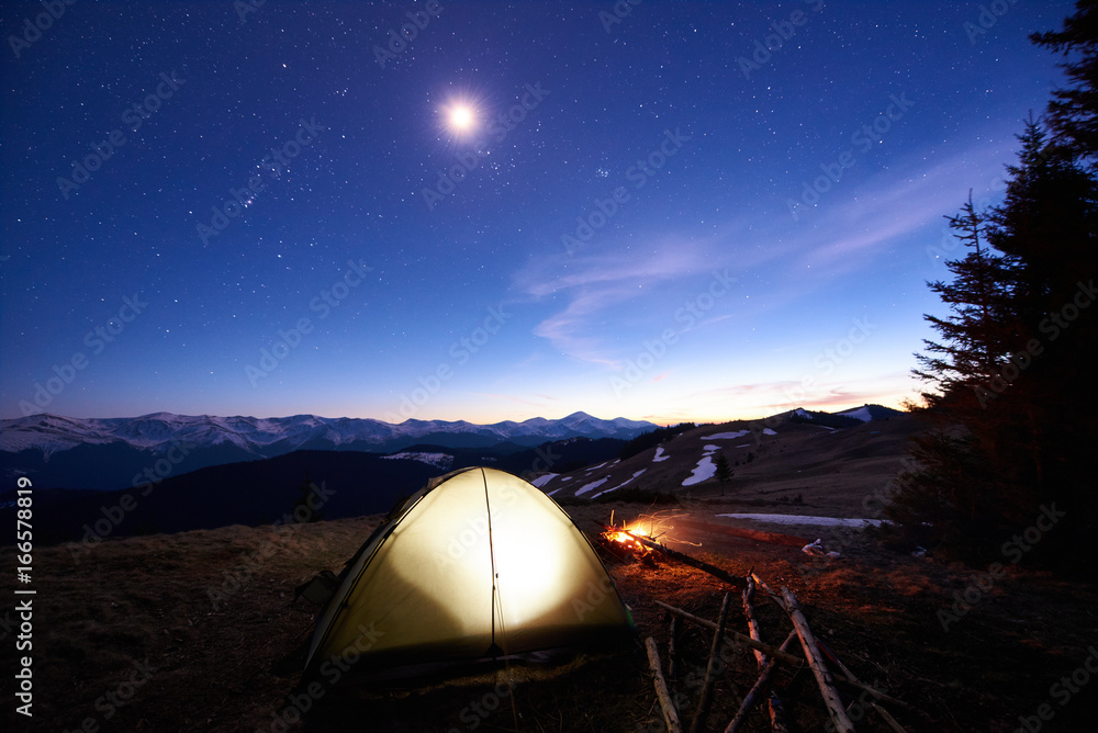 Tourist camping near forest in the mountains. Illuminated tent and campfire under beautiful evening sky full of stars and the moon