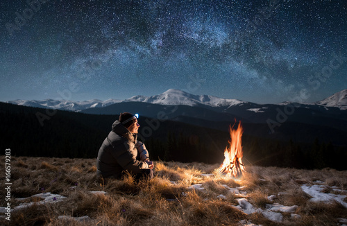 Male tourist have a rest in the mountains at night. Man with a headlamp sitting near campfire under beautiful night sky full of stars and milky way, and enjoying night scene photo